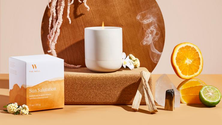 Sun salutation candle burning on a yoga block, with citrus, palo santo and flowers beside it