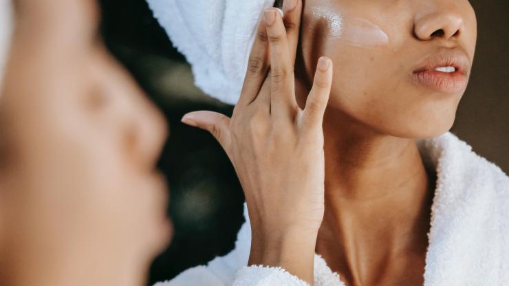 A woman dressed in a white robe applies lotion to her face.