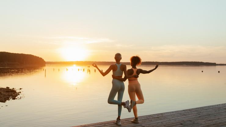 Two women hold a yoga pose while looking out at a sunset over a lake.