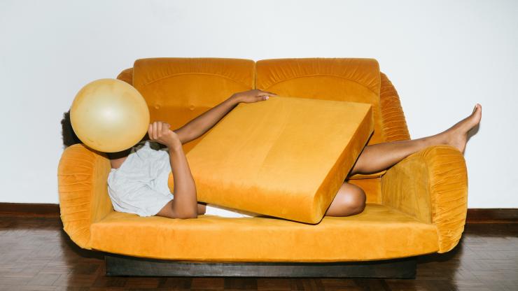 A woman lays on a bright orange couch and covers her face with an orange balloon.